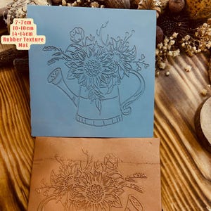 May include: A blue rubber stamp with a design of a watering can filled with sunflowers. The stamp measures 7-7cm by 10-10cm and is 14-14cm thick. The stamp is labeled "Rubber Texture Mat".