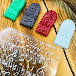 K&ouml;nnte beinhalten: Vier rechteckige, farbenfrohe Seifenst&uuml;cke mit Musikmotiven in Gr&uuml;n, Grau, Rot und Hellblau, jeweils mit einem Notenschl&uuml;ssel und Noten. Eine transparente Acrylplatte mit ge&auml;tzten Noten und Symbolen ist ebenfalls vorhanden.