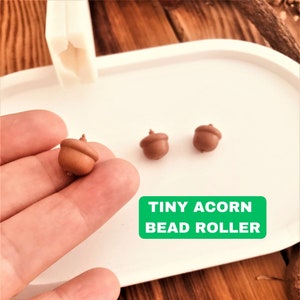 Puede incluir: Tres rodillos de cuentas de color marrón con forma de bellota sobre una superficie blanca. El texto "TINY ACORN BEAD ROLLER" está impreso en verde sobre un rectángulo blanco.