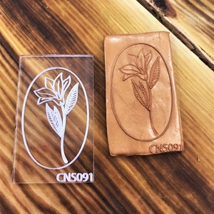 May include: A clear acrylic stamp with a floral design and the text "CNS091". The stamp is used to create an impression on leather. The impression is shown on a piece of brown leather.