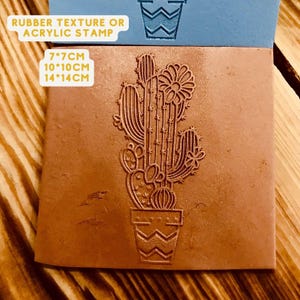 May include: A brown rubber or acrylic stamp featuring a cactus design. The cactus is in a pot with a zig-zag pattern. The stamp is available in three sizes: 7x7 cm, 10x10 cm, and 14x14 cm.
