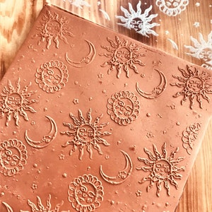 Op de afbeelding: Een close-up van een bruine kleistempel met een herhalend patroon van zonnen en manen. De stempel wordt gebruikt om een afdruk op een oppervlak te maken.