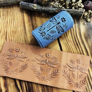 Puede incluir: Un sello cilíndrico azul con un diseño de libélula y flores se encuentra sobre un sello rectangular de color terracota con un diseño de libélula y flores. Los sellos están sobre una superficie de madera con plantas secas.