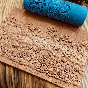May include: A blue roller with a detailed ocean-themed design rests above a rectangular, brown clay stamp. The stamp features an embossed underwater scene with waves, coral, turtles, and starfish. The roller is cylindrical and has a textured surface.
