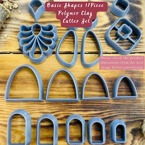 May include: A set of 17 gray polymer clay cutters in various shapes, including arches, ovals, and a leaf design. The cutters are arranged on a wooden surface. Basic Shapes 17Piece Polymer Clay Cutter Set.