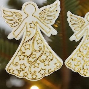 May include: Two white angel ornaments with gold accents, wings, and star patterns. The text "CHRISTMAS ANGEL STAMP+CUTTER" is at the bottom. These ornaments are likely for Christmas decorations.