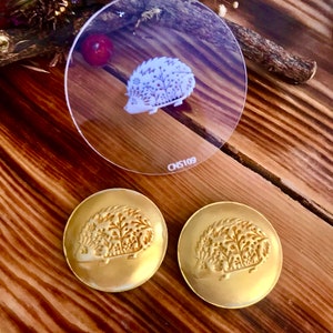 Könnte beinhalten: Zwei goldfarbene Metallknöpfe mit einem erhabenen Design eines Igels mit einem Blumenmuster. Eine durchsichtige Plastikfolie mit einem weißen Igeldesign und dem Text "CNS109" ist ebenfalls zu sehen.