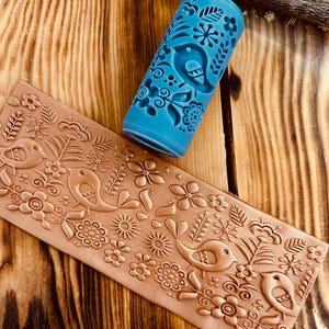 Puede incluir: Un rodillo azul con un diseño de pájaros y flores junto a una losa de arcilla marrón con el mismo patrón en relieve. El rodillo es cilíndrico y presenta un patrón repetido de pájaros, flores y hojas. La losa de arcilla es rectangular.
