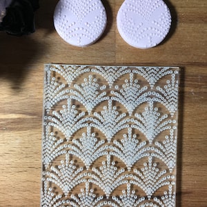 May include: Two light pink teardrop-shaped earrings with a dotted pattern, next to a clear acrylic sheet with a white dotted fan design. The items are on a wooden surface.