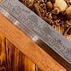 Puede incluir: Dos reglas con temática de vacaciones de invierno. Una es gris, la otra es marrón. Ambas reglas tienen un diseño grabado detallado de casas, árboles y renos. La regla gris mide hasta 30 cm.
