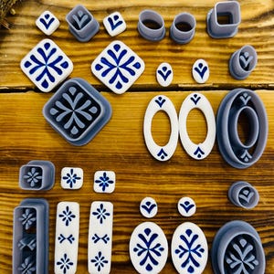 May include: A set of 19 polymer clay cutters featuring various geometric shapes and designs, including squares, ovals, and circles. The cutters are in shades of gray and white, with some featuring blue and white patterns.