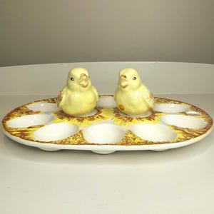 Może przedstawiać: Owalna ceramiczna podstawka na jajka z białym środkiem i obwódką z żółtych i brązowych płatków słonecznika. Na podstawce znajdują się dwie żółte ceramiczne figurki kurcząt oraz osiem okrągłych wgłębień przeznaczonych na jajka.