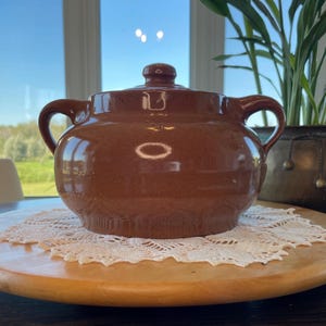 Könnte beinhalten: Eine braune Keramik-Auflaufform mit Deckel und zwei Griffen. Die Schüssel steht auf einem Serviertablett aus Holz mit einer weißen Spitzendecke. Eine Topfpflanze befindet sich im Hintergrund.