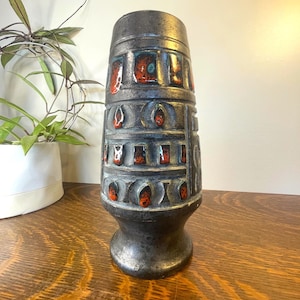 May include: A tall, cylindrical ceramic vase with a dark, matte charcoal-grey finish. The surface features a repeating geometric pattern of recessed rectangular and arched panels, each accented with a glossy, vibrant orange and black lava glaze.