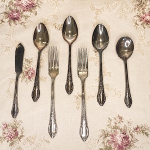 Puede incluir: Un juego de seis utensilios de metal plateado con detalles florales en relieve en los mangos. La colección incluye un cuchillo para mantequilla, dos tenedores, dos cucharas grandes y una cuchara pequeña, dispuestos en una fila vertical.