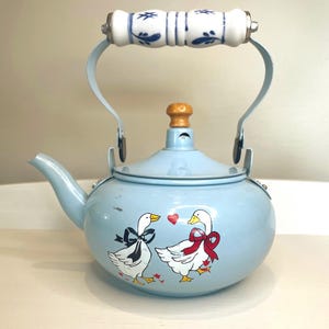 May include: A light blue metal tea kettle featuring a graphic of two white geese with red and black bows and a small red heart. The kettle has a white ceramic handle with blue floral patterns and a small wooden knob on the lid.