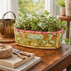 Puede incluir: Una jardinera rectangular de cerámica con una planta verde y blanca. La jardinera tiene un diseño decorativo de hojas en tonos verdes, amarillos y marrones. Una pequeña regadera de latón se encuentra en el fondo.