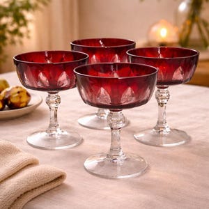 May include: Four ruby red champagne coupe glasses with clear stems and bases are arranged on a light pink tablecloth. The glasses have a faceted design on the bowls. A plate of fruit and a folded beige napkin are in the background.