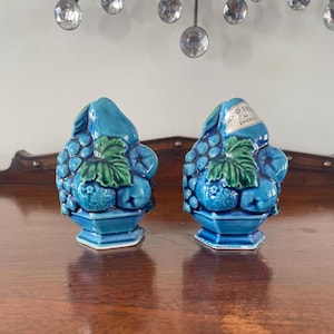 Może przedstawiać: Dwie turkusowe ceramiczne figurki w kształcie owoców. Każda figurka przedstawia kiść winogron, gruszkę i zielone liście. Jedna figurka ma etykietę z napisem "Mood Indigo by INARCO". Figurki znajdują się na drewnianej powierzchni.