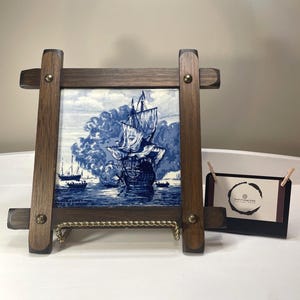 May include: A wooden picture frame with a blue and white nautical scene tile. The frame has a dark wood finish and brass accents. A small black card holder with a business card is to the right.