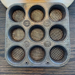 Puede incluir: Un molde para magdalenas de metal vintage con una superficie texturizada y ocho moldes individuales para magdalenas. El molde está ligeramente oxidado y tiene las palabras "Ovenised Made in England" impresas en dos de los moldes.