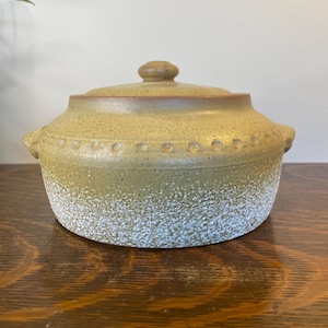 Peut inclure: Une cocotte en céramique ronde avec couvercle, présentant une partie supérieure beige mouchetée et une base blanche texturée. Le récipient comprend deux petites poignées latérales et une rangée d'indentations circulaires sur le bord.