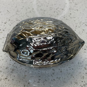 Può includere: Un contenitore argentato a forma di noce, con apertura a cerniera. Il contenitore ha una superficie strutturata che imita le creste di un guscio di noce. Il contenitore è chiuso e riflette la luce, suggerendo una finitura metallica lucida.