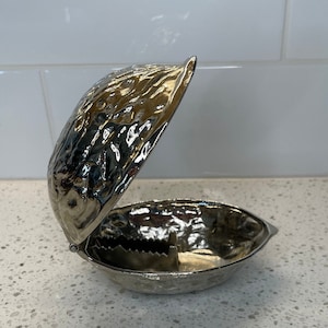 May include: A silver-coloured nutcracker shaped like a walnut, open to show the cracking mechanism. The surface has a textured, realistic walnut shell appearance. The nutcracker is on a white surface.