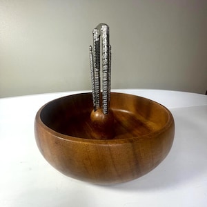 May include: A wooden bowl with a silver nutcracker. The bowl is made of a dark brown wood and has a smooth, rounded shape. The nutcracker is silver and has a decorative design. The nutcracker is placed in the center of the bowl.
