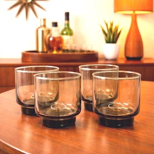 May include: Four clear, lowball glasses with a dark grey base and rim, arranged on a wooden table. The glasses have a simple, modern design. A bar cart with bottles and a lamp are in the background.