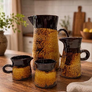 May include: A ceramic set consisting of one large pitcher, one smaller pitcher, and two mugs. Each piece features a dark brown glazed top section and a textured, mottled golden-yellow and brown base with a distinct crackled, porous appearance.