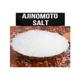 Op de afbeelding: Een close-up van een kom met witte Ajinomoto-zoutkristallen op een houten oppervlak. Het label luidt "Ajinomoto Salt".
