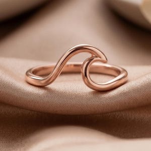 Puede incluir: Un anillo de oro rosa con una banda continua que se eleva formando una onda suave y estilizada en la parte superior. El metal tiene un acabado pulido y reflectante, y el diseño consiste en una línea fluida que forma la cresta de la ola.