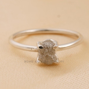 Pode incluir: Um anel de prata com um diamante cinza bruto como peça central. O diamante tem uma forma natural e irregular. A banda do anel é fina e polida, criando um design minimalista. O anel é apresentado sobre um fundo neutro.