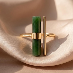 Op de afbeelding: Een goudkleurige ring met een verticale, zeshoekige donkergroene stenen staaf in het midden, gecombineerd met een parallelle, slanke goudkleurige metalen staaf. Het ontwerp is open en toont een gepolijste afwerking en een gladde steen.