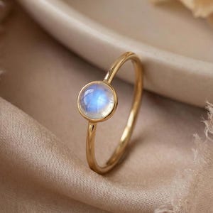 Puede incluir: Un anillo de oro con una piedra preciosa redonda de color azul claro. El anillo está engastado en una banda delgada y se muestra sobre una superficie de tonos neutros. La piedra preciosa tiene un brillo sutil e iridiscente.