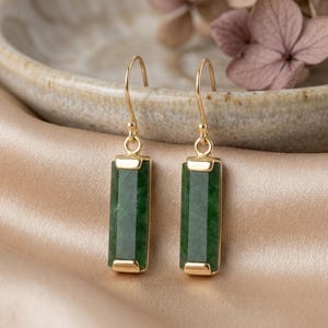 May include: A pair of gold-colored drop earrings with rectangular, faceted, dark green jade stones. The earrings hang from a simple hook design. The background is a soft, golden-colored fabric.