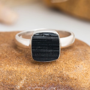 Pode incluir: Um anel de prata com uma pedra preta retangular. A banda do anel é lisa e polida, e a pedra preciosa tem uma superfície texturizada. O anel está sobre uma superfície castanha.