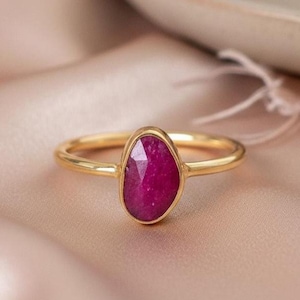 Op de afbeelding: Een gouden ring met een facetgeslepen, ovaalvormige, dieproze edelsteen. De ring ligt op een zachte, perzikroze stof. De wazige achtergrond toont een decoratief element en een keramische schaal.