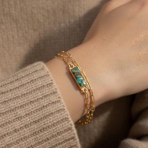 Könnte beinhalten: Goldfarbenes Armband mit einem rechteckigen Türkisstein in der Mitte. Das Armband hat ein Doppelketten-Design und wird am Handgelenk getragen. Der Hintergrund ist ein neutrales Beige. Der Türkisstein ist ca. 1,5 cm lang.