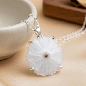 Puede incluir: Collar con cadena plateada y colgante blanco y redondo. El colgante presenta un diseño radial y un pequeño centro marrón. El collar se muestra sobre una superficie beige, con un cuenco blanco y una bandeja de madera en el fondo.