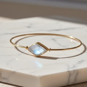 Op de afbeelding: Een gouden armband met een ruitvormige, iriserende edelsteen. De edelsteen heeft blauwe en witte tinten en is gezet in een gouden bezel. De armband wordt gepresenteerd op een wit marmeren oppervlak.