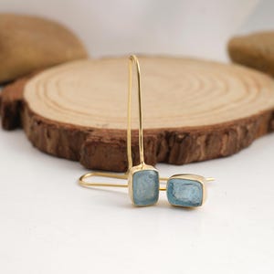 Puede incluir: Un par de pendientes colgantes dorados con piedras preciosas rectangulares de color azul claro. Los pendientes se muestran sobre una superficie blanca con un fondo de madera. Las piedras est&aacute;n engastadas en un bisel dorado.
