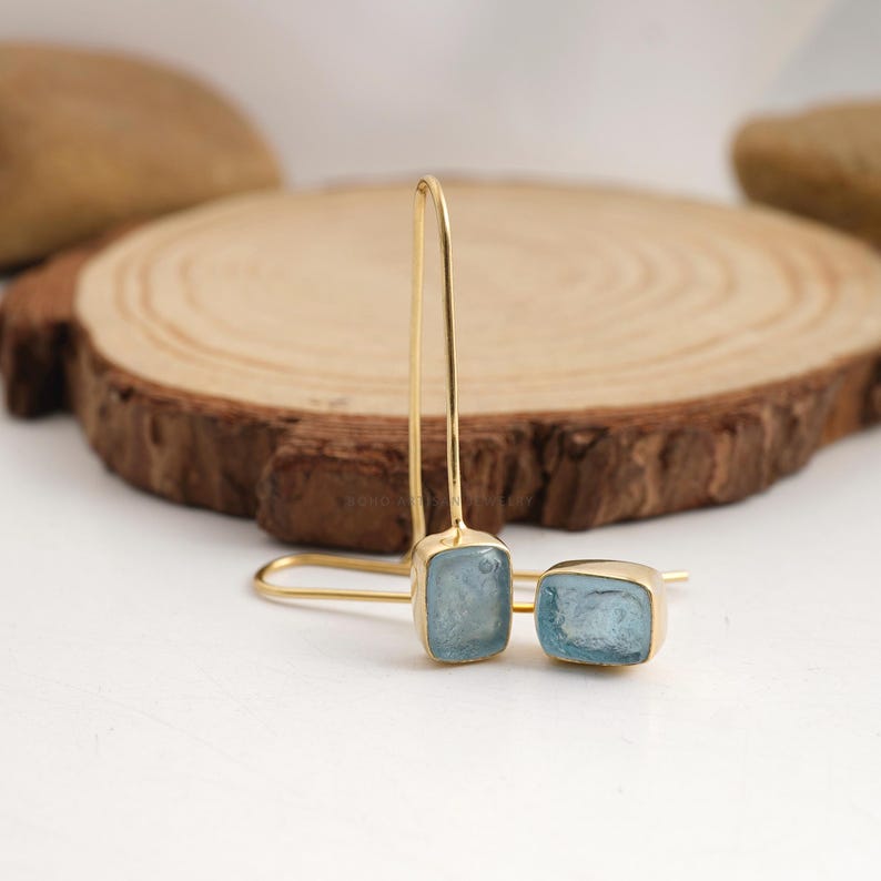 Puede incluir: Un par de pendientes colgantes dorados con piedras preciosas rectangulares de color azul claro. Los pendientes se muestran sobre una superficie blanca con un fondo de madera. Las piedras est&aacute;n engastadas en un bisel dorado.