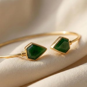 May include: A gold-toned bracelet with two emerald-green, geometric-shaped gemstones. The bracelet has an open design, with the gemstones at each end. The gemstones are set in gold and have a faceted appearance.