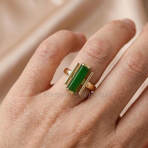 May include: A gold-colored ring featuring a rectangular, vibrant green stone. Two parallel gold bars flank the stone, adding a modern touch. The ring is worn on a finger, showcasing the jewelry's design. The text "BOHO ARTISAN JEWELRY" is visible.