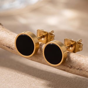May include: A pair of stud earrings featuring a circular design with a smooth, flat black center inlay surrounded by a polished gold-tone metal rim. The earrings include standard post backs and are displayed resting on a light-colored wooden surface.