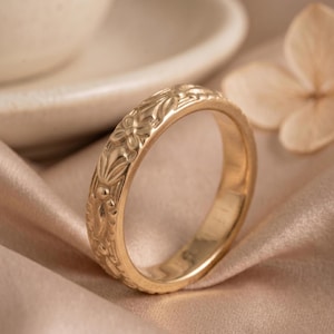 Puede incluir: Un anillo de metal en tono dorado que presenta un patrón floral en relieve continuo alrededor de la banda exterior. El anillo tiene un acabado pulido y un perfil redondeado, mostrando un diseño repetitivo de pequeñas flores y hojas grabadas.