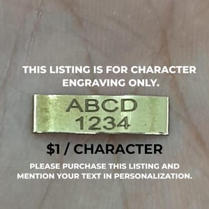 Pode incluir: Uma pequena etiqueta retangular de metal dourado com o texto gravado "ABCD 1234". A etiqueta é mostrada com o texto "THIS LISTING IS FOR CHARACTER ENGRAVING ONLY."