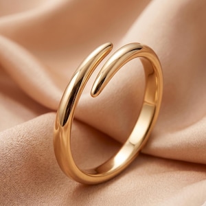 Puede incluir: Un anillo abierto de tono dorado pulido con una banda lisa y redondeada que se estrecha en los extremos. El diseño es de estilo bypass con dos terminales puntiagudos ligeramente superpuestos, creando una superficie metálica continua.